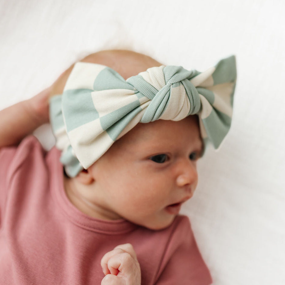 A close-up lifestyle shot of a baby wearing a large, knotted bow headband in sage green and cream, paired with a dusty rose pink top, lying on a soft white blanket.