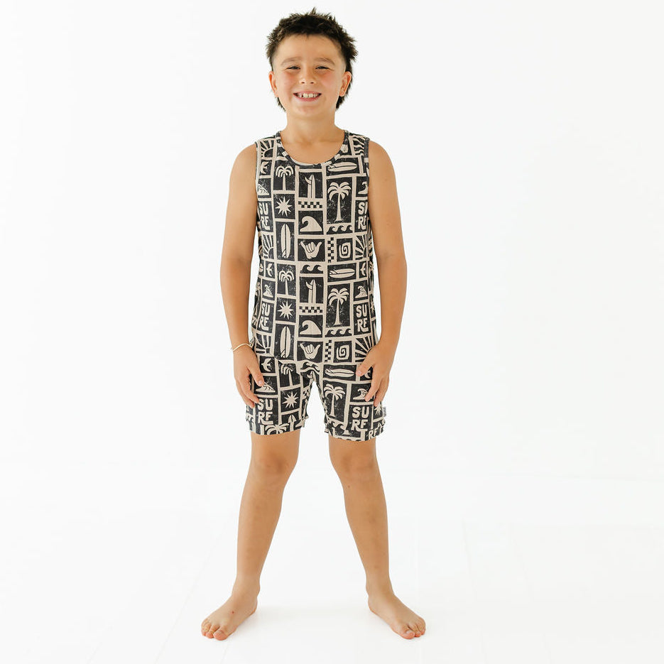 A young boy stands barefoot against a plain white studio background, wearing a sleeveless black and white patterned top and matching shorts.