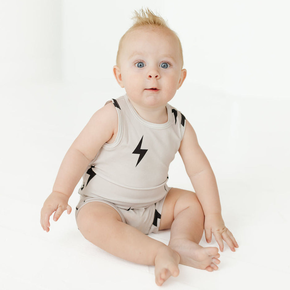A baby sitting on a white studio floor wearing a light beige sleeveless romper with a black lightning bolt graphic on the chest.