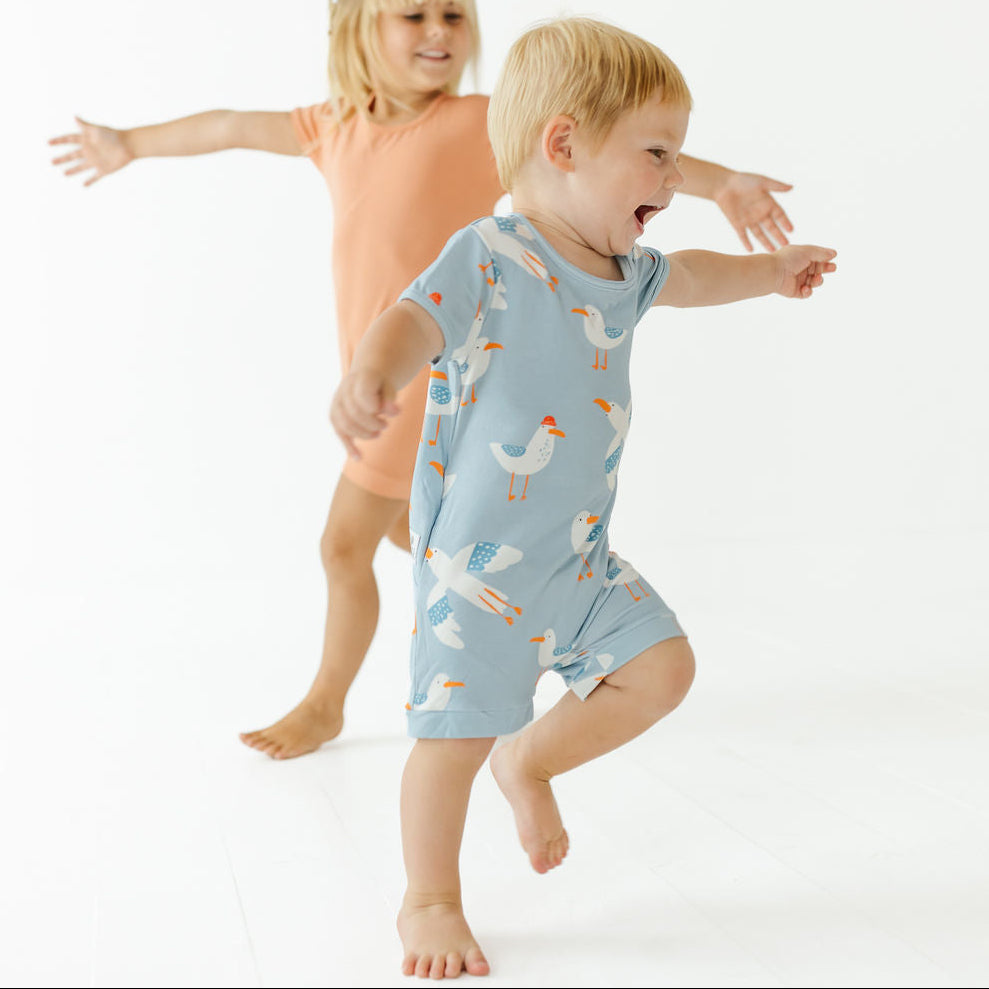 A toddler in a light blue seagull-print romper jumps barefoot in front of a girl in an orange dress, set against a seamless white studio background.