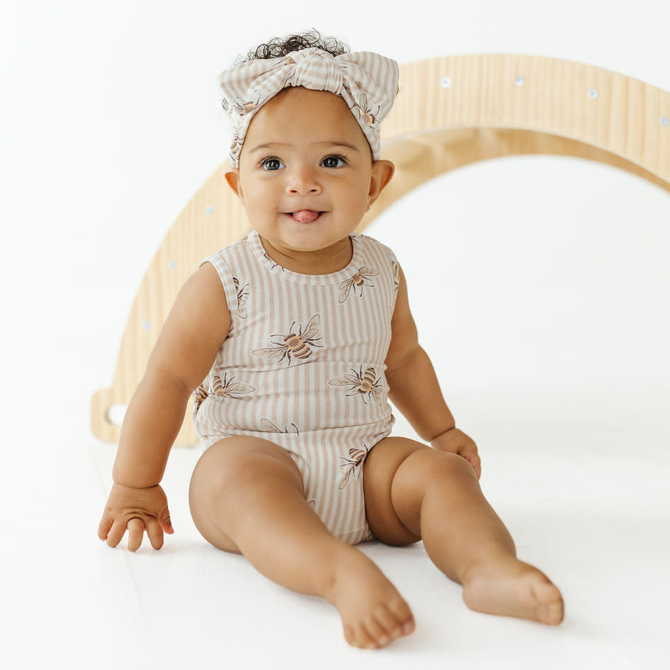 A baby sitting on a white studio floor wearing a sleeveless beige and white striped romper with embroidered bees and a matching striped bow headband, posed in front of a light wood arch.