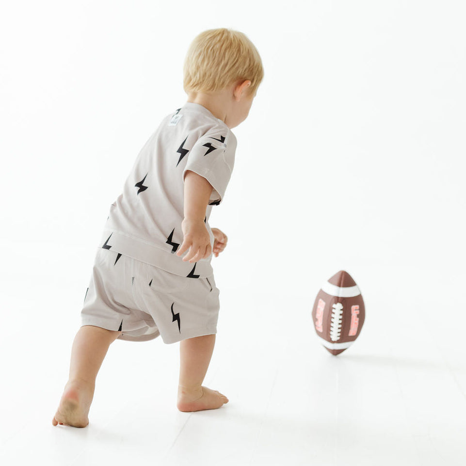 A toddler wearing a light gray short-sleeve romper with black lightning bolt prints is seen from behind, reaching toward a small brown football on a clean white studio background.