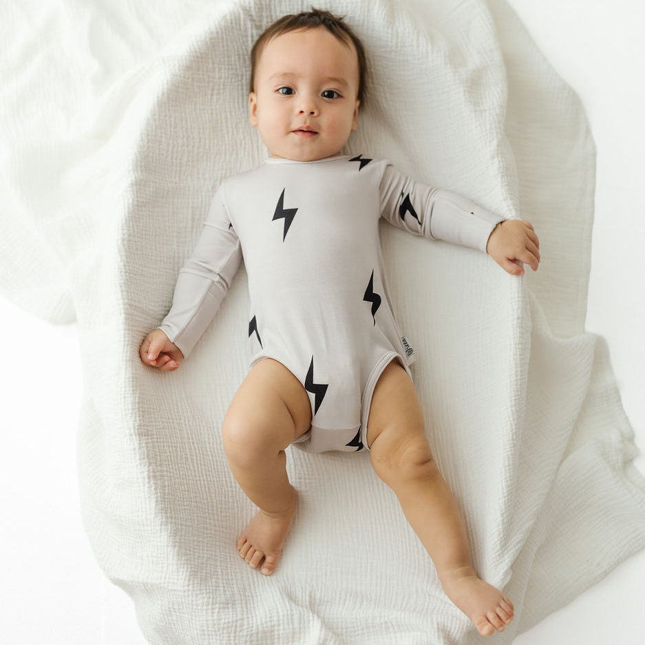 A baby lying on a white muslin blanket, wearing a light gray long-sleeve bodysuit with black lightning bolt prints.