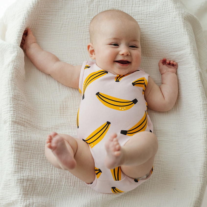 A baby lying on a textured cream muslin blanket, wearing a pale pink sleeveless bodysuit with a yellow banana print.