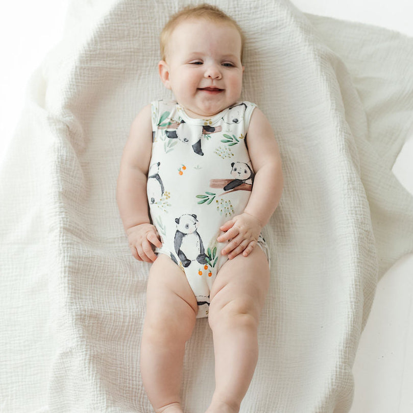 A baby lying on a textured white muslin blanket, wearing a sleeveless bodysuit with a cute panda and nature print.
