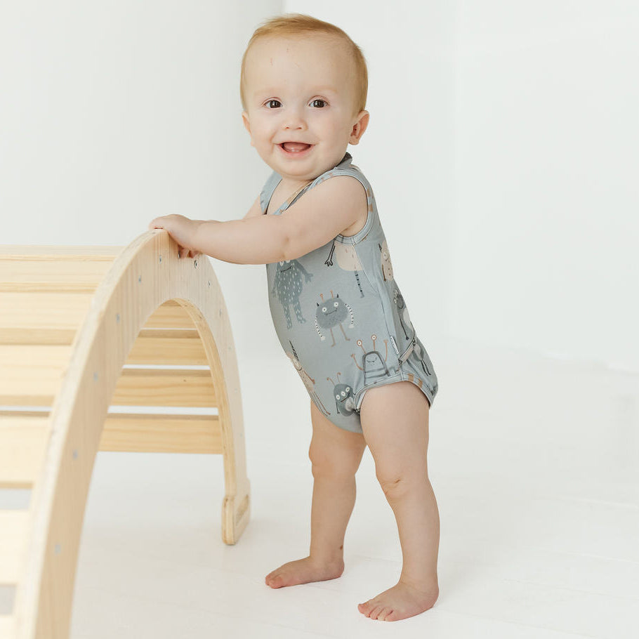 A baby standing on a white studio floor and holding onto a light wood arch play structure, wearing a sleeveless light gray-blue romper with a cat print.