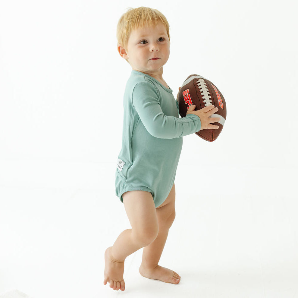 A toddler standing barefoot on a white studio background, wearing a long-sleeve sage green romper and holding a brown American football.