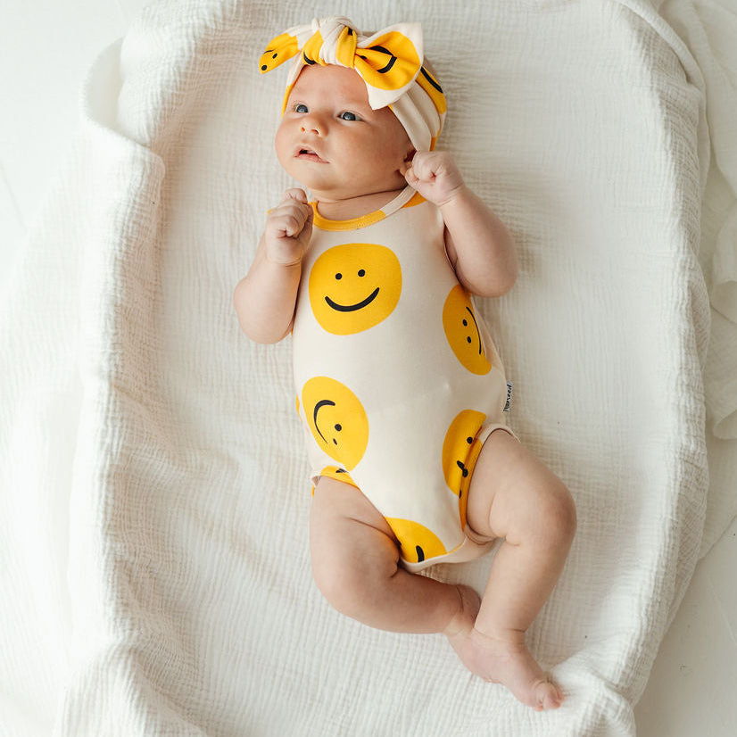 A baby lying on a white textured blanket, wearing a cream bodysuit with yellow smiley faces and a matching bow headband.