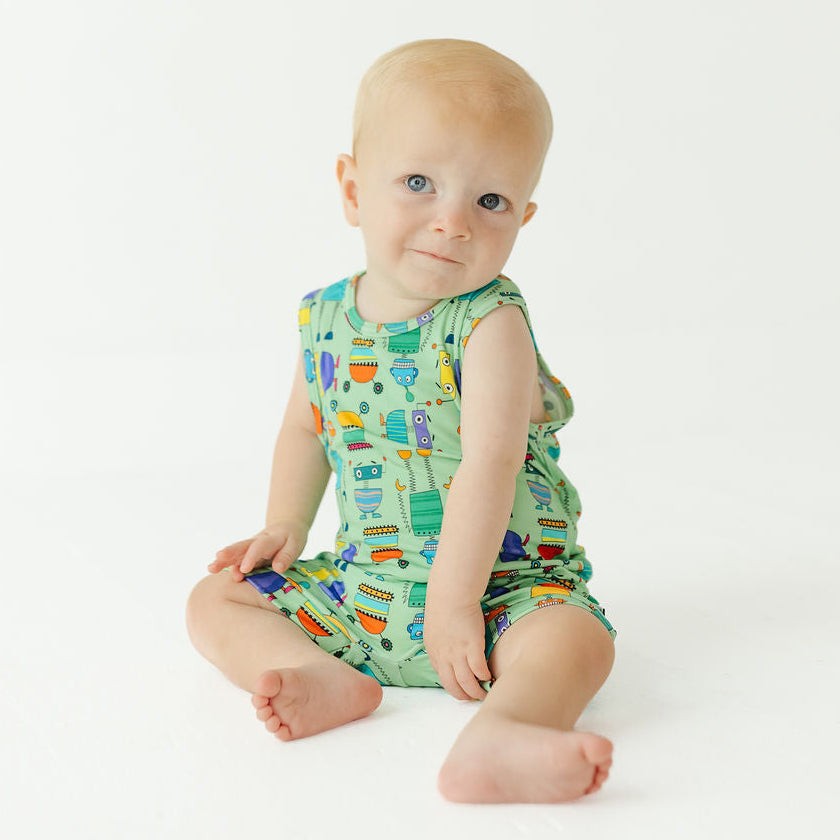 A baby sitting on a white studio background wearing a green sleeveless romper with a colorful construction tool pattern.