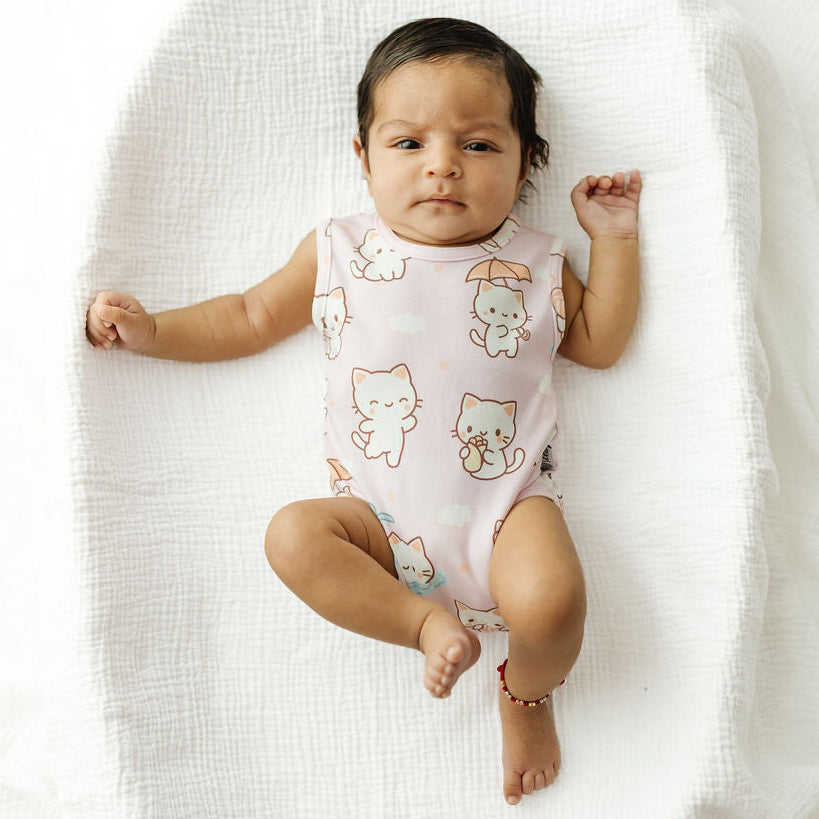 A baby lying on a white muslin bassinet wearing a light pink sleeveless bodysuit with a white cat print.