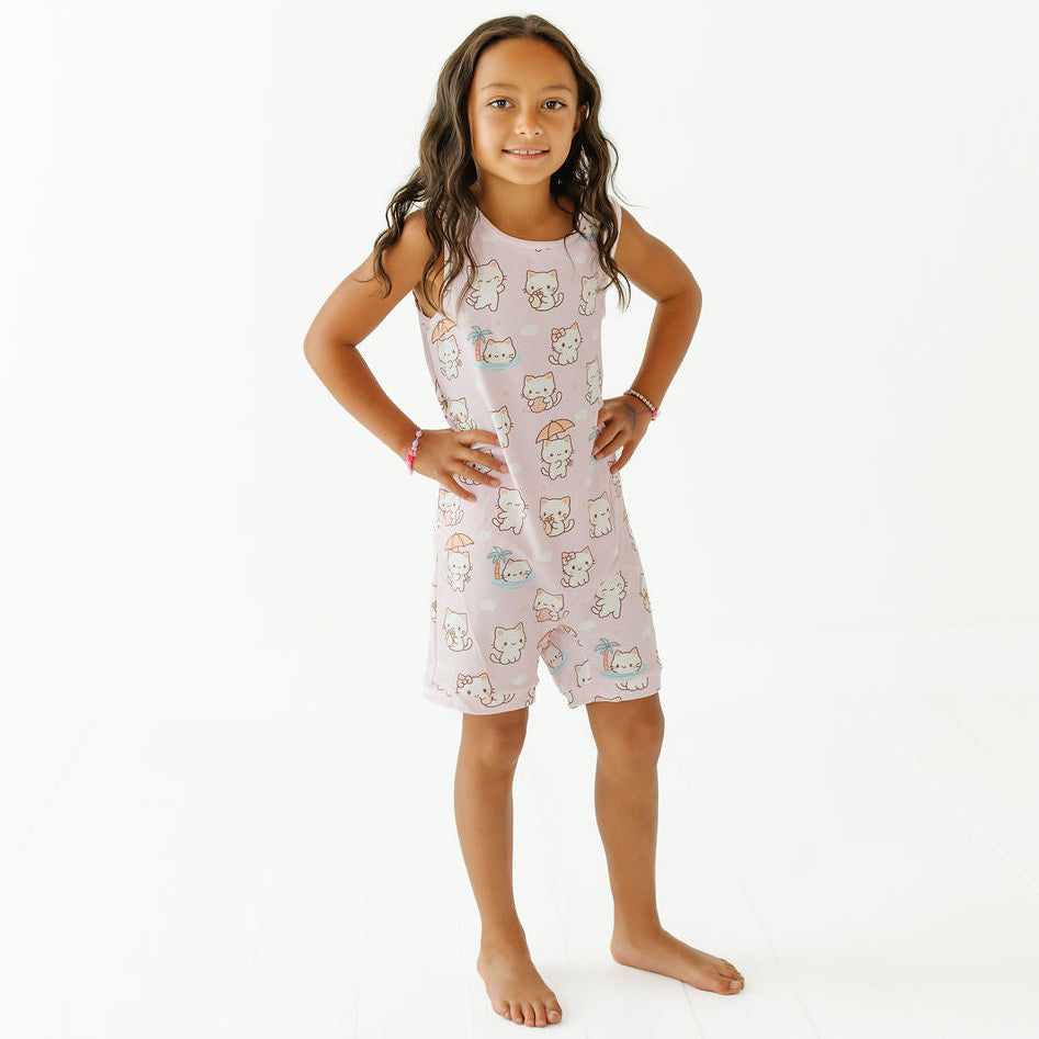 A young girl stands barefoot against a plain white studio background, wearing a sleeveless light pink romper or pajama set with an allover Hello Kitty character print.