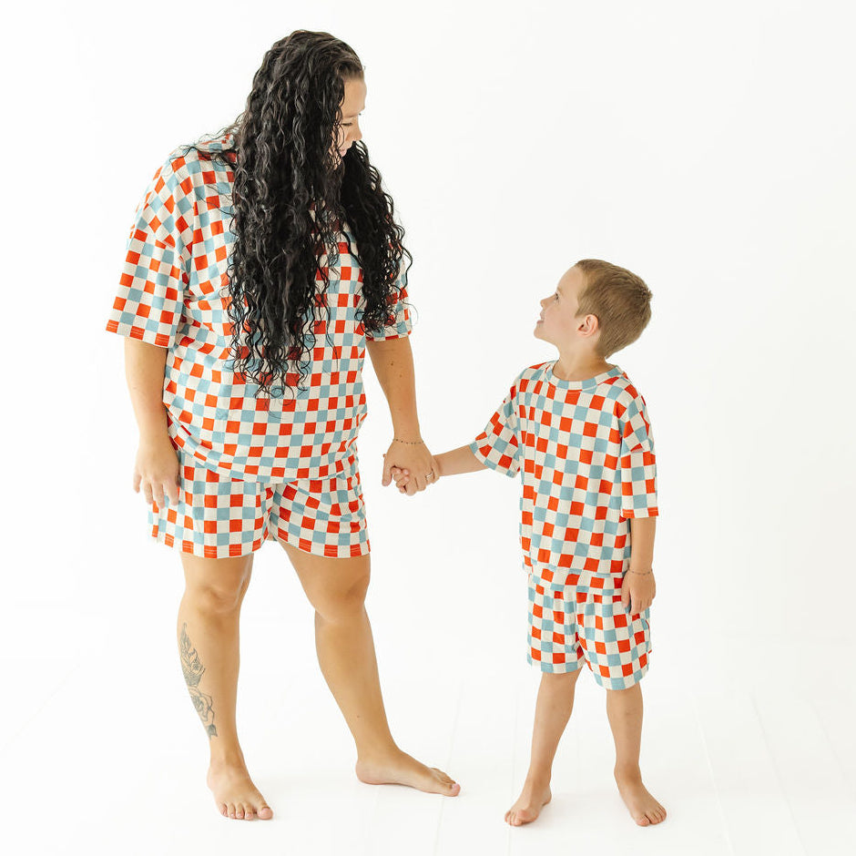 A woman and a young boy wearing matching short-sleeve pajama sets with a red, orange, and white checkerboard pattern, holding hands against a plain white background.