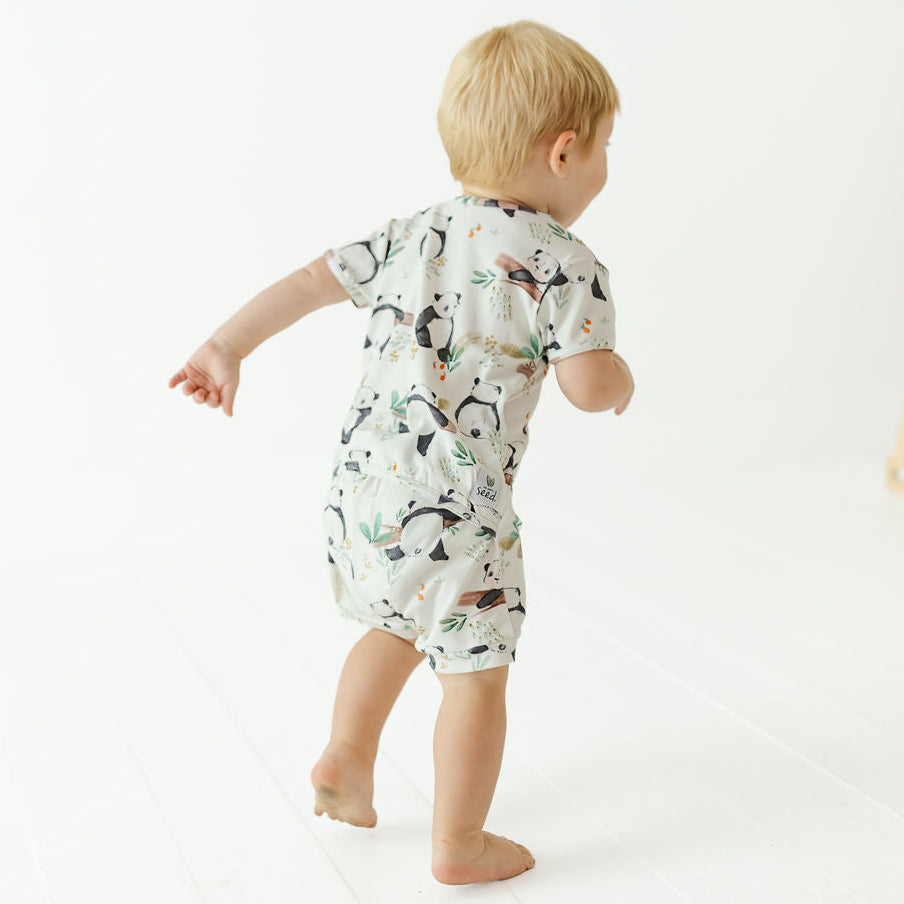 A toddler with blond hair seen from behind, wearing a short-sleeve romper with a black and white panda print and walking barefoot on a white floor.