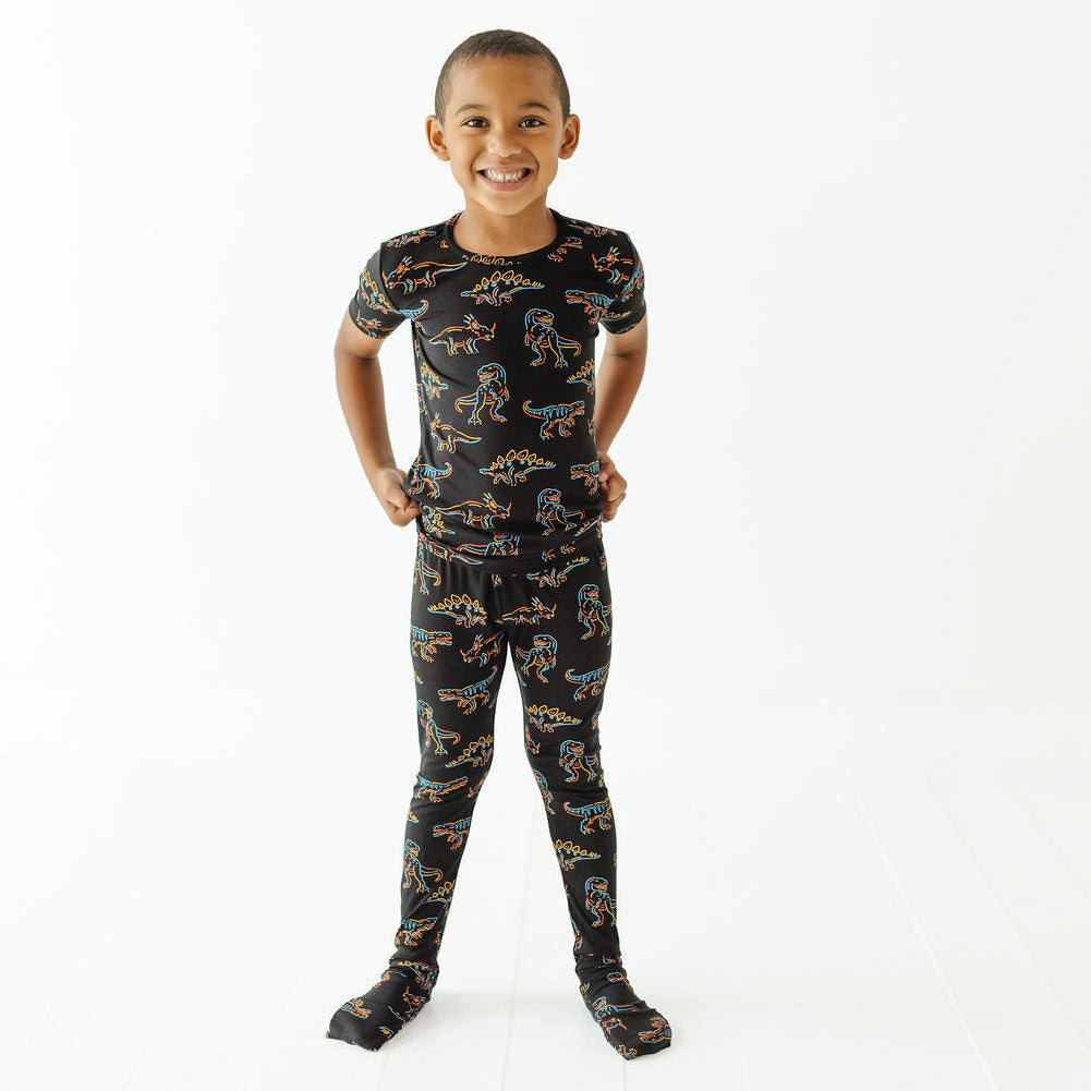 A young boy smiling and posing in a matching black pajama set with a colorful dinosaur print, consisting of a short-sleeve top and footed pants, against a plain white studio background.