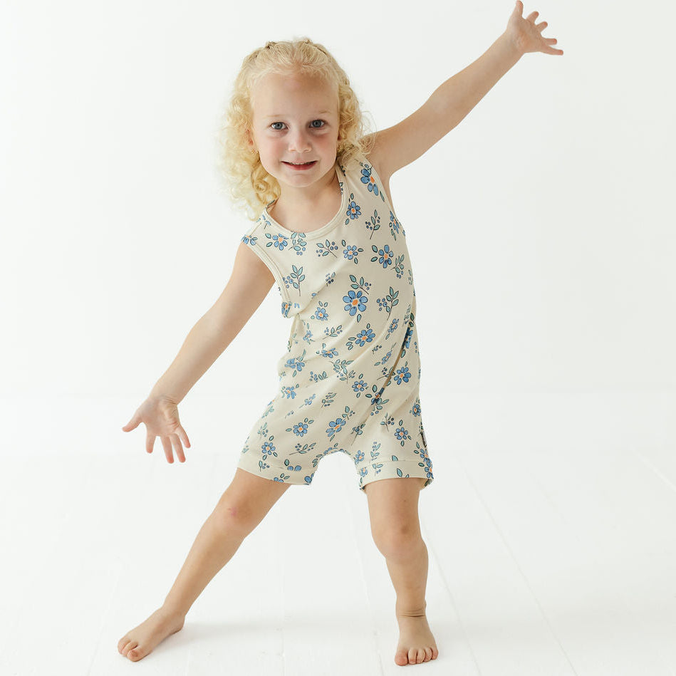 A young blonde girl posing in a sleeveless cream-colored romper with a blue floral pattern against a bright white studio background.