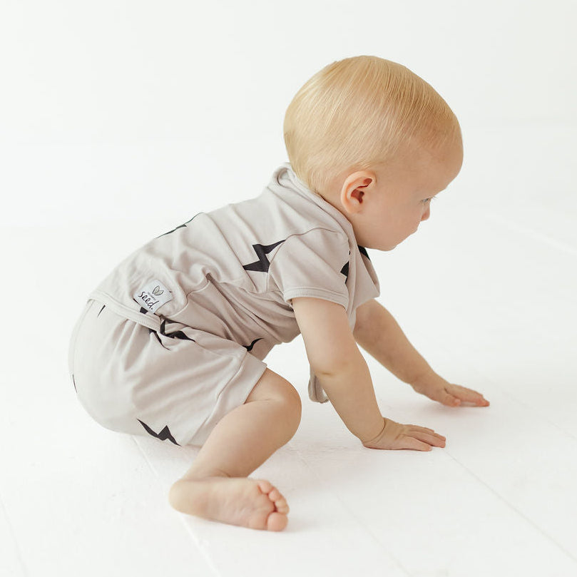 A baby crawling on a white wooden floor, wearing a light beige short-sleeve romper with black lightning bolt prints. The brand name 'Kings of NY' is visible on a small patch on the back of the romper.