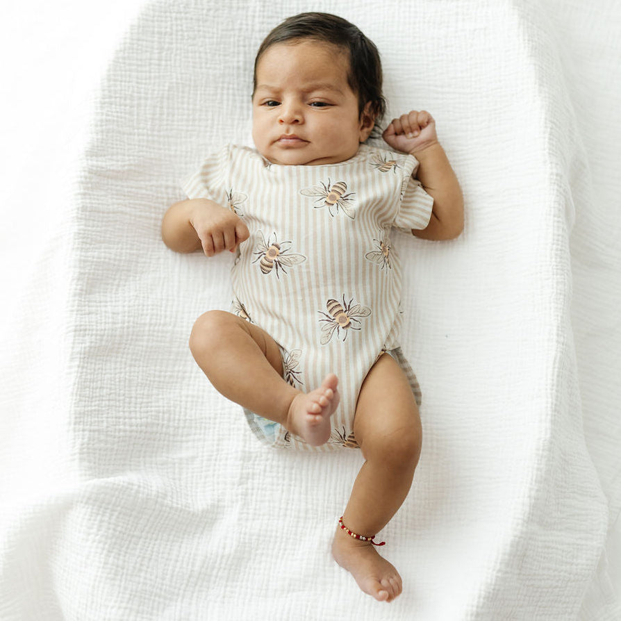 A baby lying on a white muslin blanket, wearing a short-sleeve cream bodysuit with a subtle vertical stripe pattern and small bee prints.