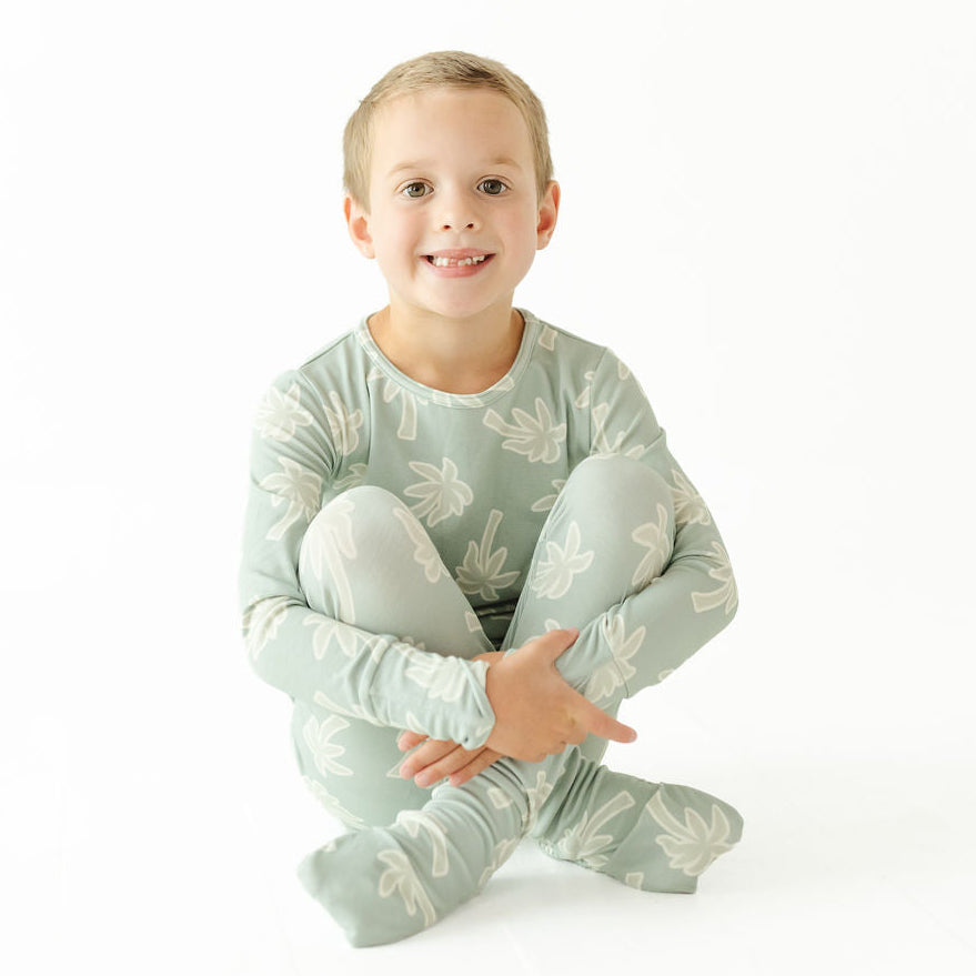 A young child sitting cross-legged on a white studio background, wearing a long-sleeve footed pajama set in a pale sage green color with a white leaf pattern.