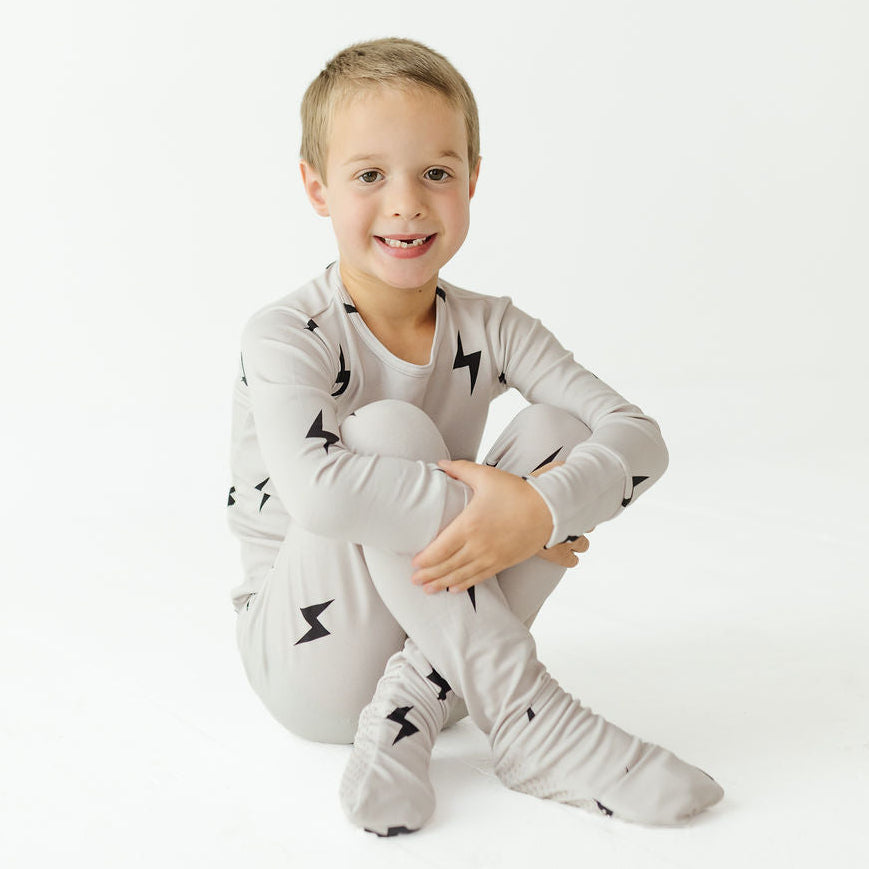 A young boy sitting cross-legged on a white studio background, wearing a light gray long-sleeve pajama set with black lightning bolt prints.
