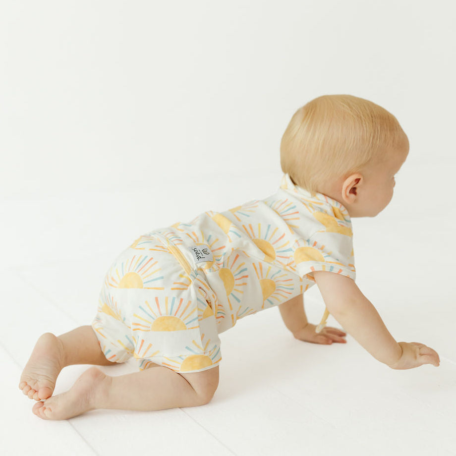 A baby crawling on a white studio floor, wearing a short-sleeve romper with a yellow sun and light blue cloud pattern on a white background.