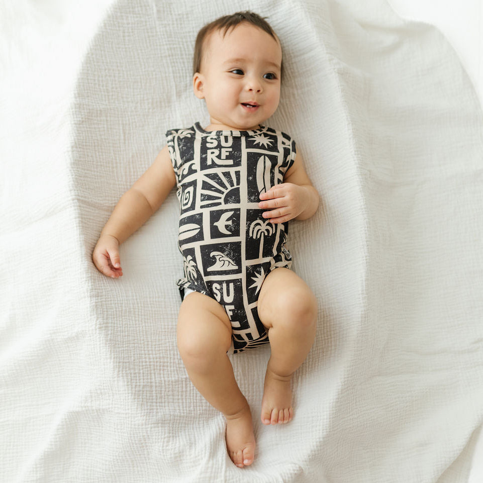 A baby lying on a white textured blanket, wearing a sleeveless black and white patterned bodysuit with surf-inspired graphics and text.
