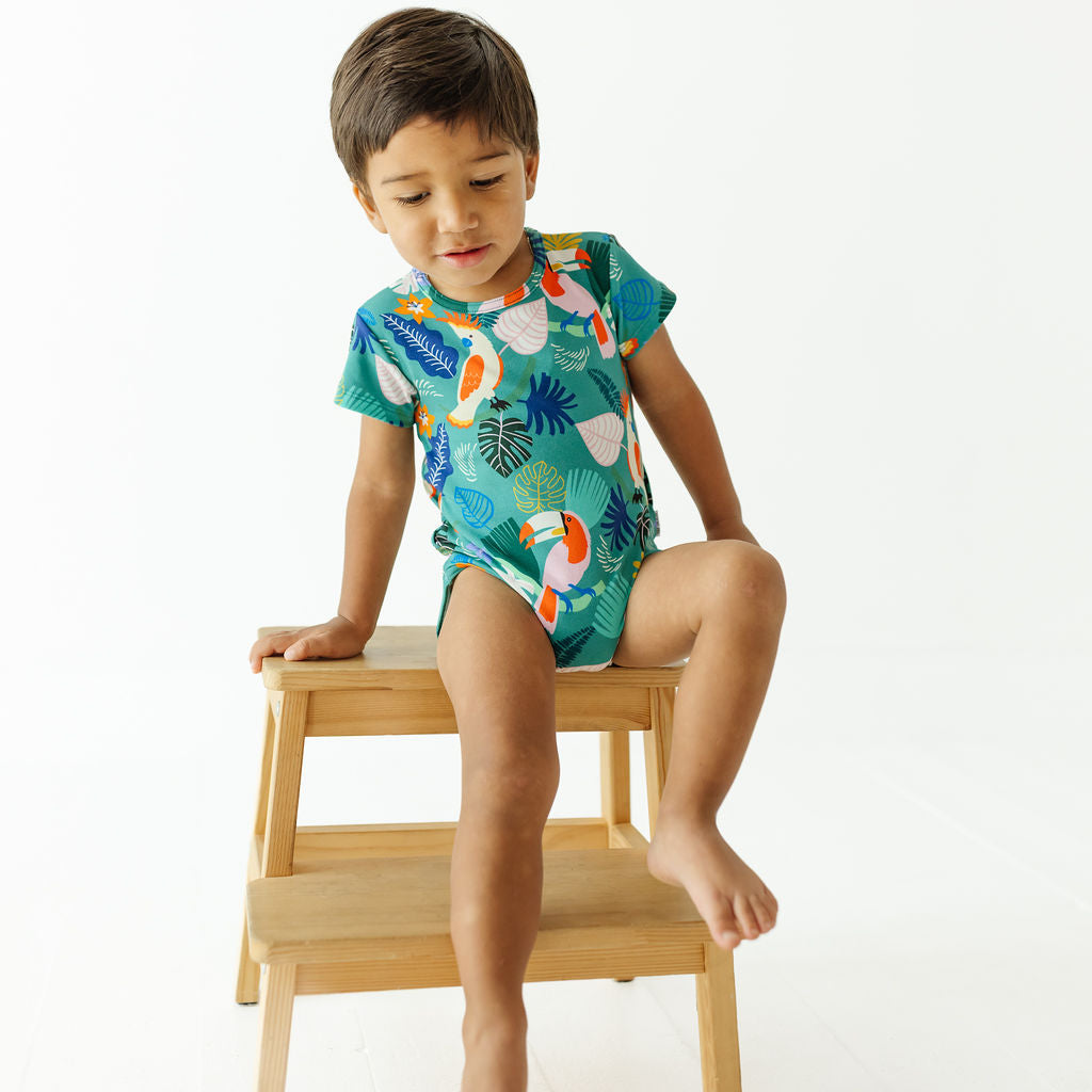 A young boy wearing a short-sleeve tropical-print baby bodysuit sits on a small wooden step stool against a clean white background.
