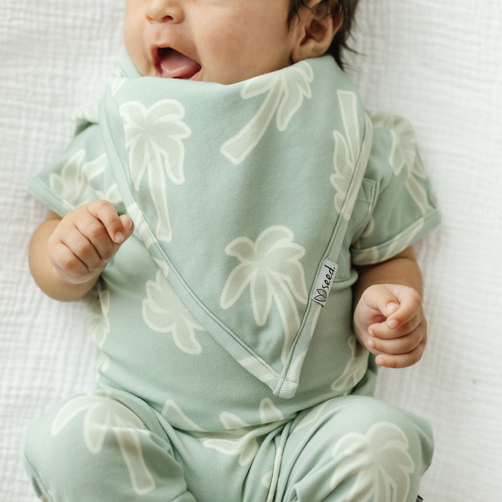 A close-up of a baby wearing a matching pale sage green short-sleeve one-piece outfit and a triangular bib with a white palm tree pattern, lying on a textured white blanket.