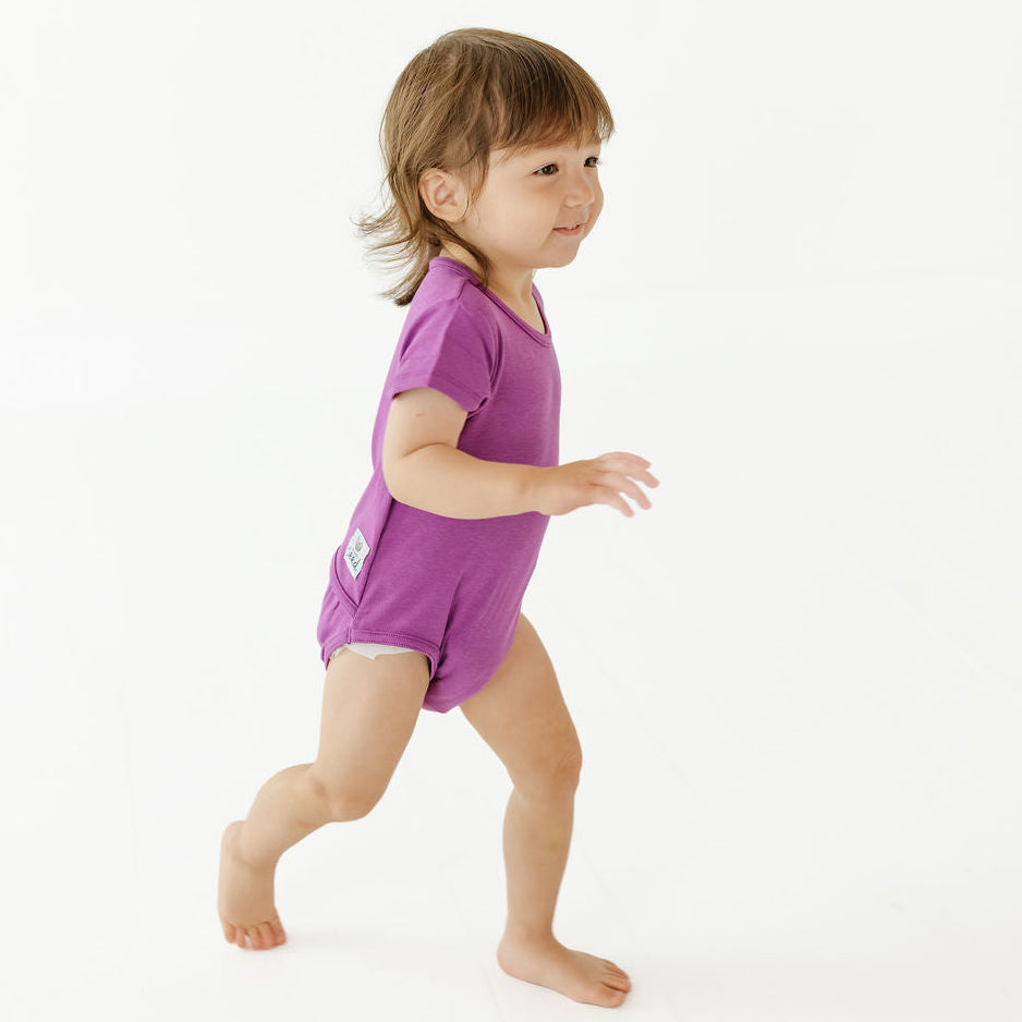 A toddler girl walking barefoot in profile, wearing a short-sleeve purple romper or bodysuit against a clean white studio background.