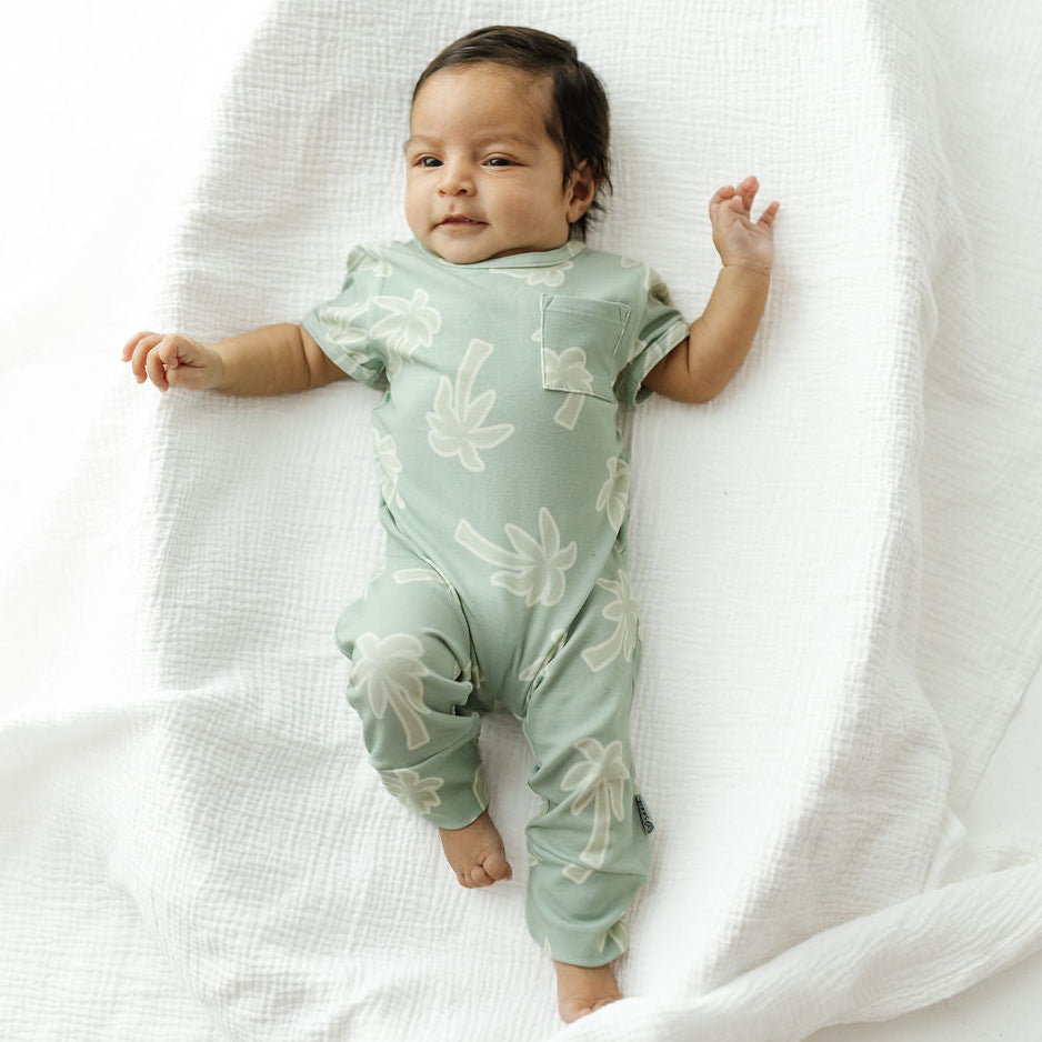 A baby lying on a white textured muslin blanket, wearing a sage green short-sleeve one-piece romper with a white palm tree print.