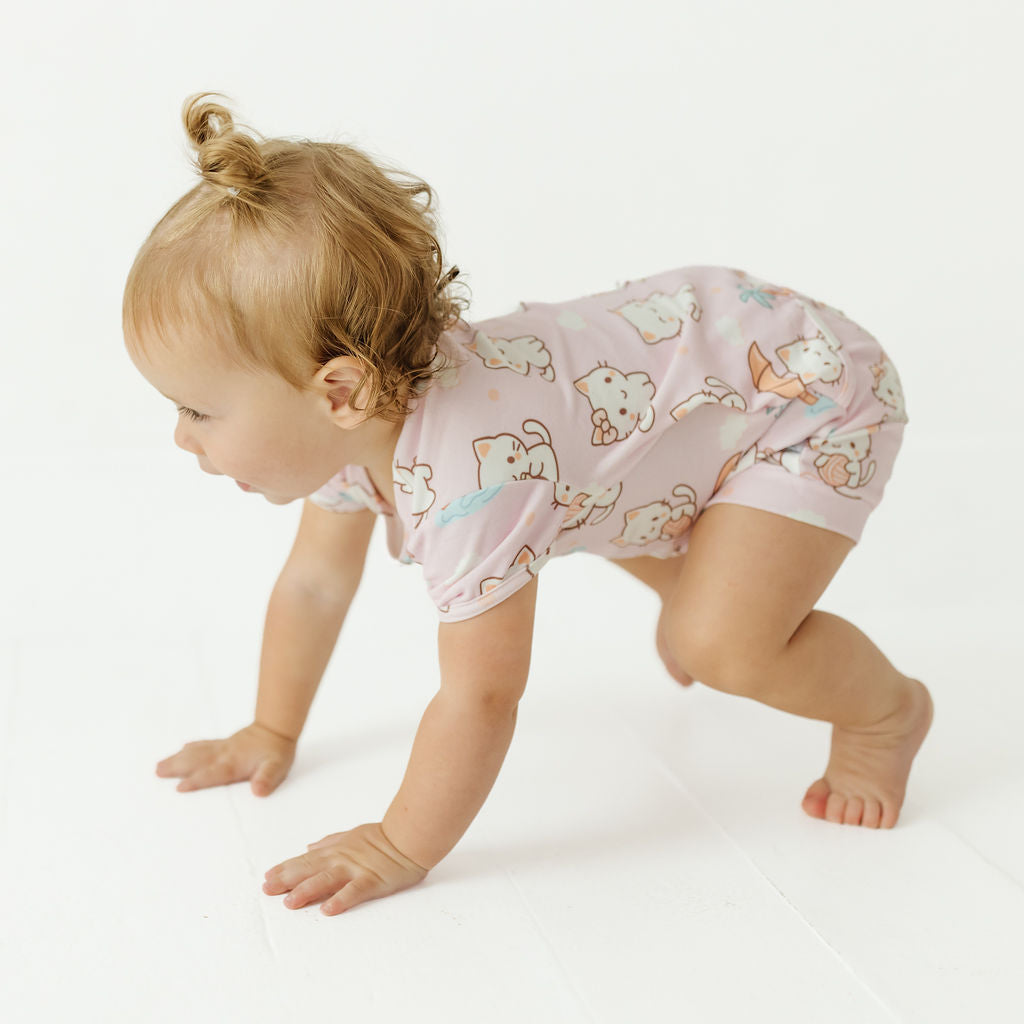 A baby crawling on a white studio floor wearing a light pink short-sleeve romper with a playful cartoon animal print.