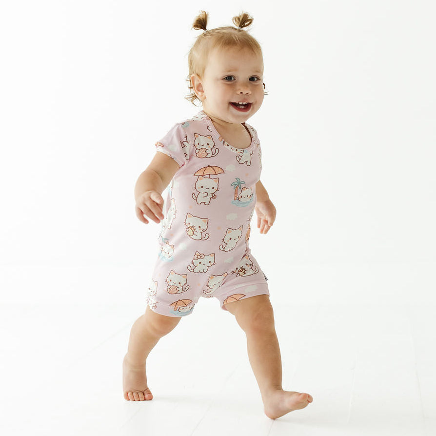 A smiling toddler walking barefoot on a clean white studio background, wearing a light pink short-sleeve romper covered in a cute cartoon cat print.