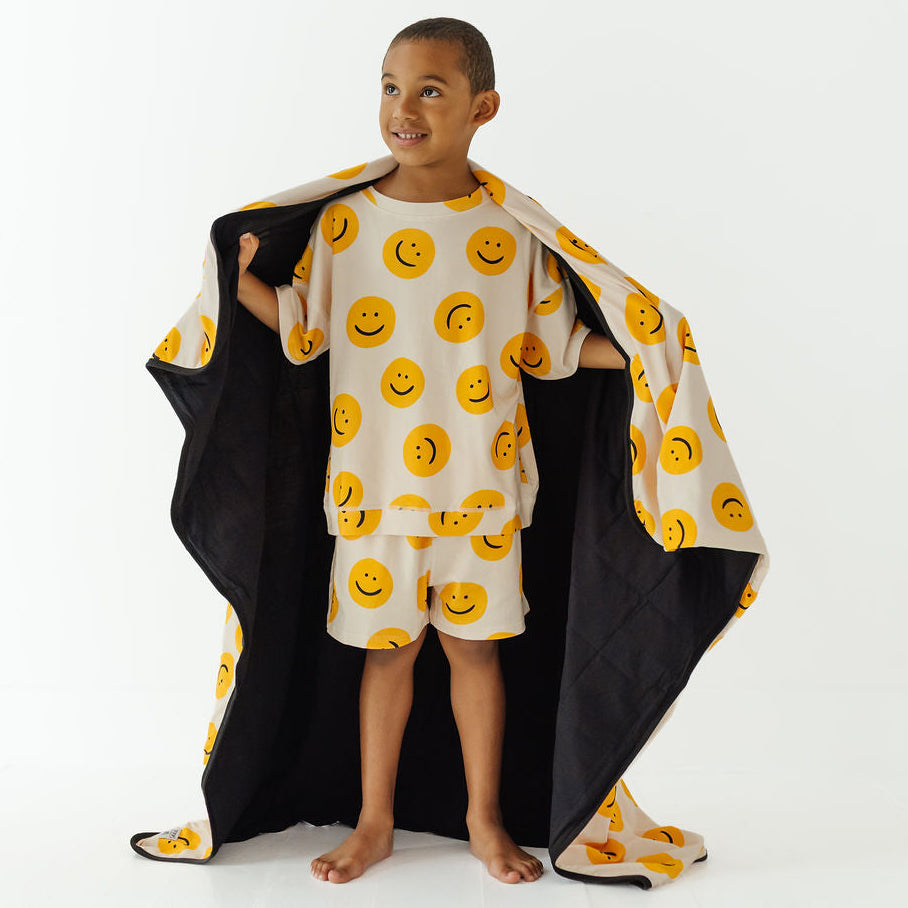 A young boy stands barefoot in a studio wearing a cream-colored pajama set with a yellow smiley face pattern, holding open a matching black quilted blanket or cape with yellow smiley faces.