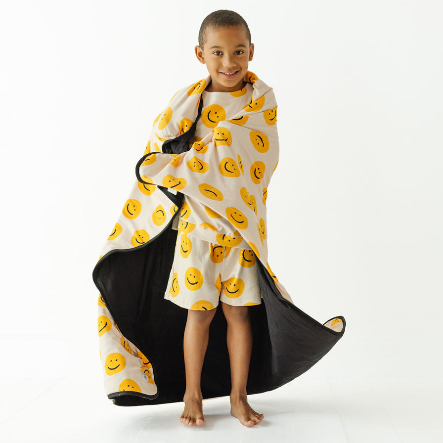 A young child standing barefoot on a white studio background, wrapped in a hooded blanket or towel that is white with a yellow smiley face pattern on the outside and black on the inside.