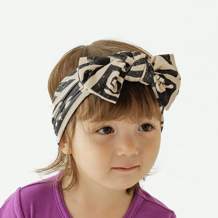 A toddler girl wearing a solid purple short-sleeve bodysuit and a black and beige patterned headband, standing against a plain white background.