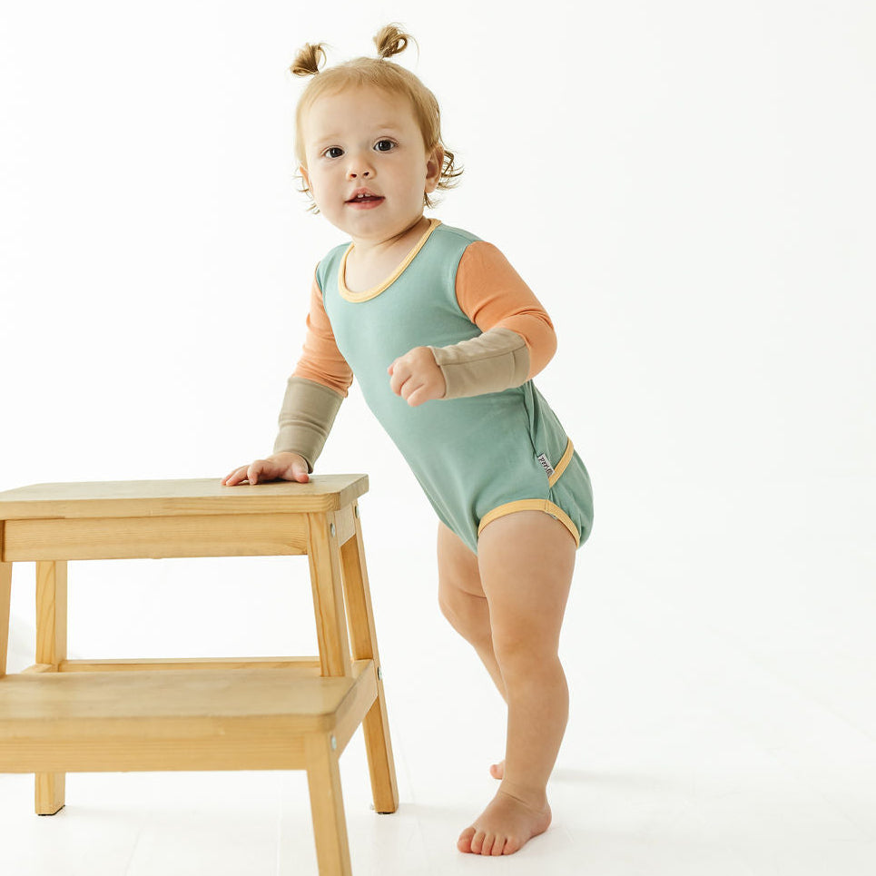 A toddler stands barefoot on a white studio floor, leaning on a small wooden step stool. The child is wearing a sleeveless sage green bodysuit with orange long sleeves and beige arm warmers.