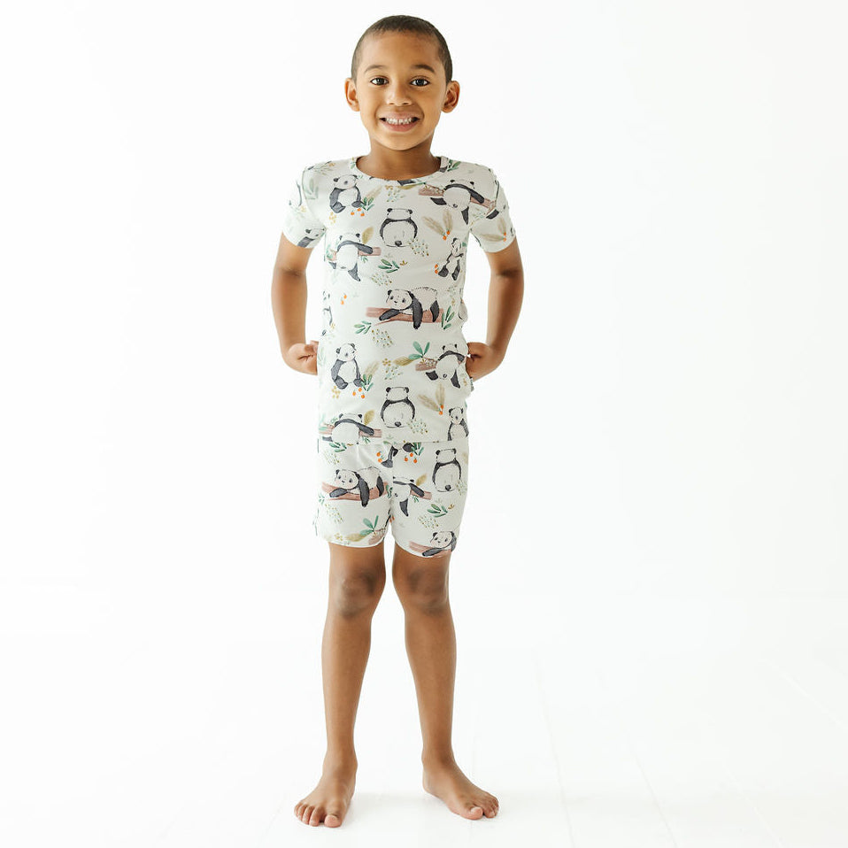 A young child standing against a plain white studio background, wearing a short-sleeve one-piece pajama romper with an allover panda print.