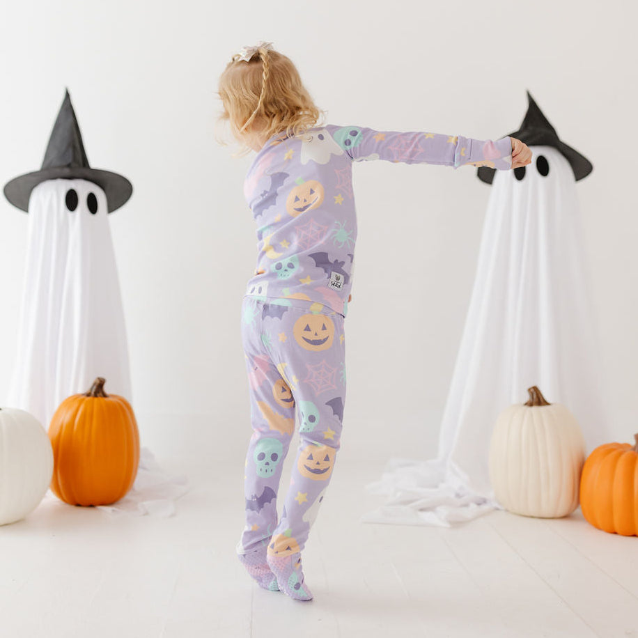 A young child wearing a light purple Halloween-themed pajama set with a pumpkin and ghost print, standing in a white studio decorated with ghost props and pumpkins.