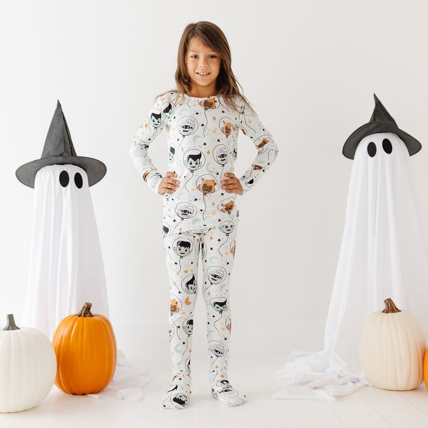 A young girl stands in a white studio wearing a white Halloween pajama set with a black cat print. She is flanked by two white ghost props wearing black witch hats, with orange and white pumpkins on the floor.