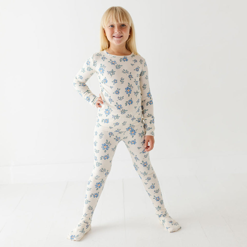 A young girl with blonde hair stands in a studio wearing a long-sleeve, full-body footed pajama suit with a blue floral pattern on a cream background.