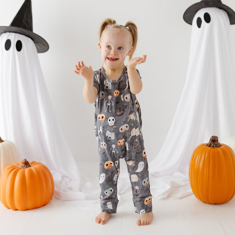 A toddler stands barefoot in a Halloween-themed studio setup, wearing a gray one-piece jumpsuit covered in cute ghost and pumpkin prints. Two large white ghost cloths with black witch hats flank the child, with orange and white pumpkins on the floor around them.