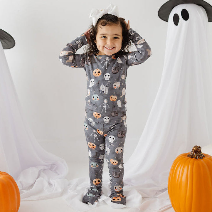 A young child wearing a gray long-sleeve pajama set with a white skull and ghost pattern stands in the center, flanked by two large white ghost decorations with black witch hats and orange pumpkins on a white studio background.