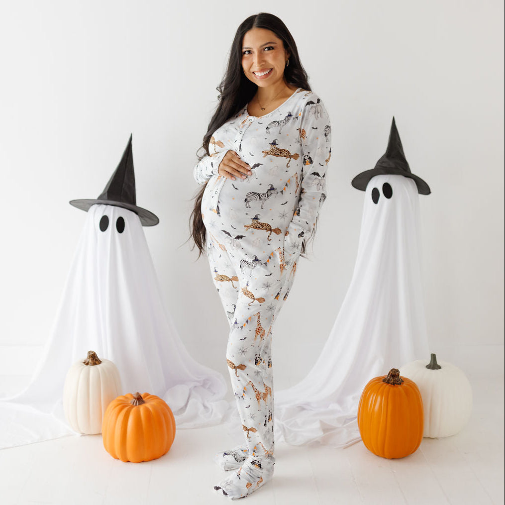 A pregnant woman stands in a Halloween-themed studio setting wearing a white long-sleeve pajama set with an all-over witch and cat print. She is flanked by two ghost decorations with black witch hats, orange and white pumpkins, and a clean white background.