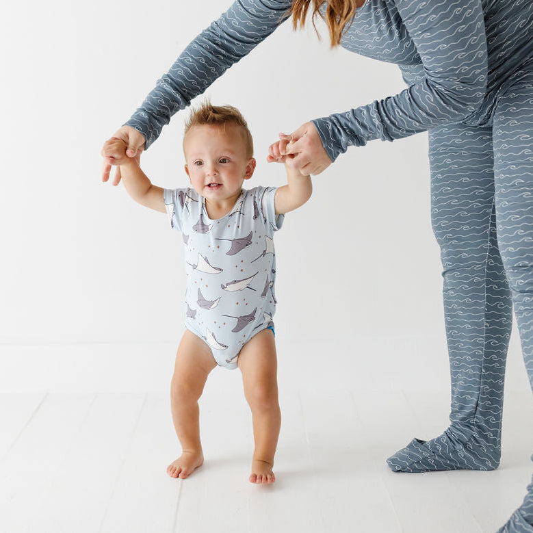 A woman and a baby wearing matching patterned sleepwear in a bright white studio. The woman wears a slate blue long-sleeve pajama set with a wavy pattern, and the baby wears a white bodysuit with a gray shark print while learning to walk.