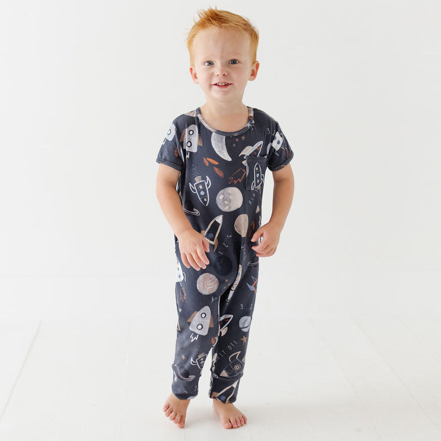 A young child with red hair stands barefoot in a white studio setting, wearing a dark navy one-piece pajama or romper covered in a space-themed print of rockets, planets, and stars.