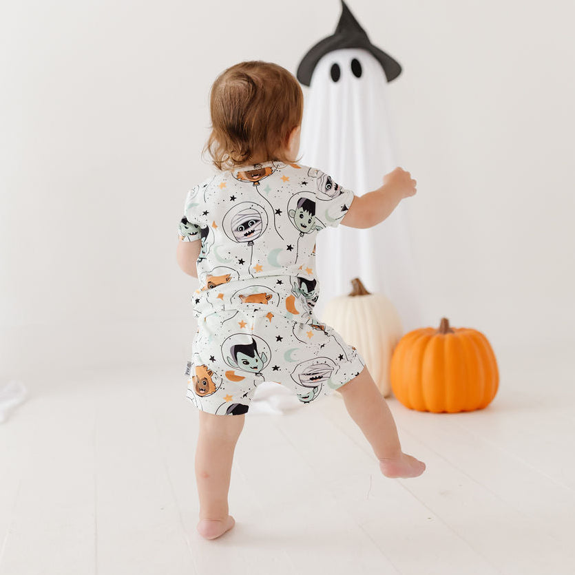 A toddler stands with their back to the camera in a white Halloween-themed room, wearing a short-sleeve romper with a cartoon astronaut and space-themed print. Decorative ghost figures, a white pumpkin, and an orange pumpkin are visible in the background.