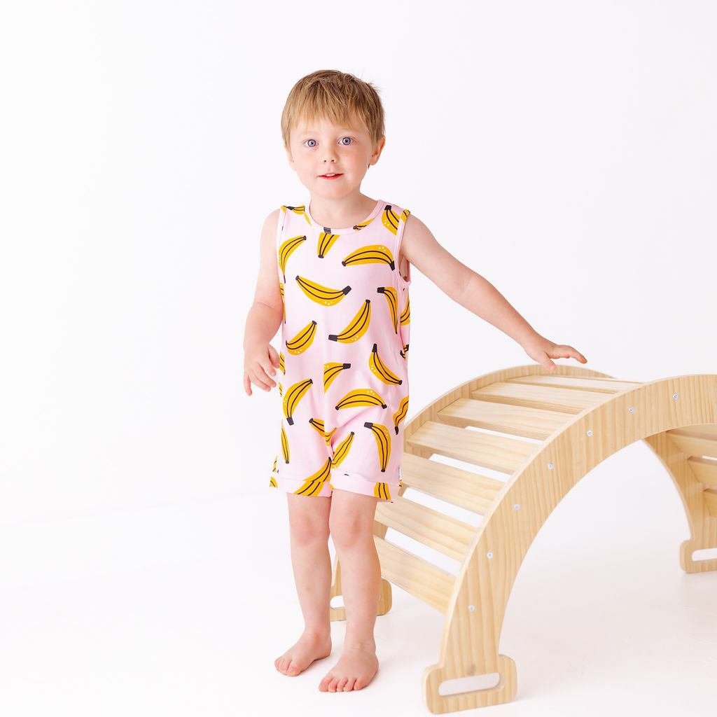 Child wearing a pink sleeveless dress with banana prints standing next to a wooden climbing toy on a white background