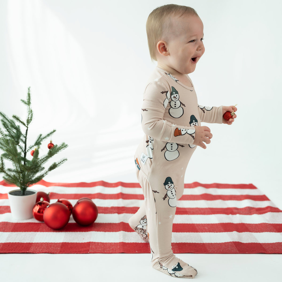 A baby wearing a beige footed pajama with a snowman print stands on a red and white striped blanket next to a small potted tree and red ornaments.