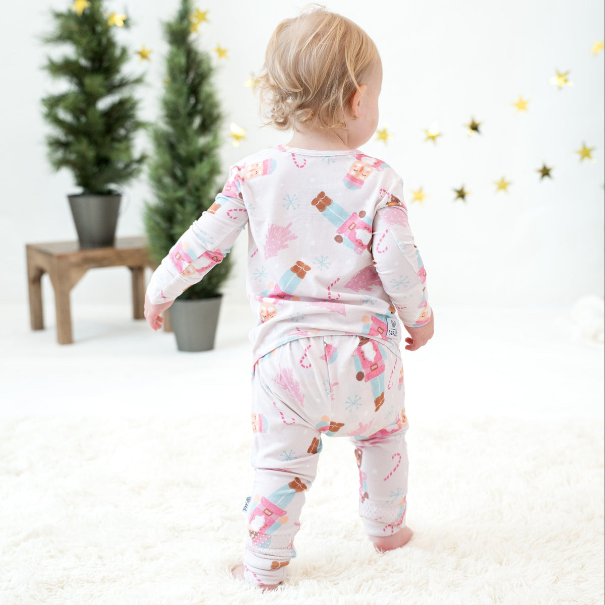A toddler standing on a white rug, viewed from behind, wearing a light pink long-sleeve pajama set with a colorful Christmas-themed print of Santa Claus, candy canes, and snowflakes. The background is a bright, minimalist room with potted trees and gold star decorations.