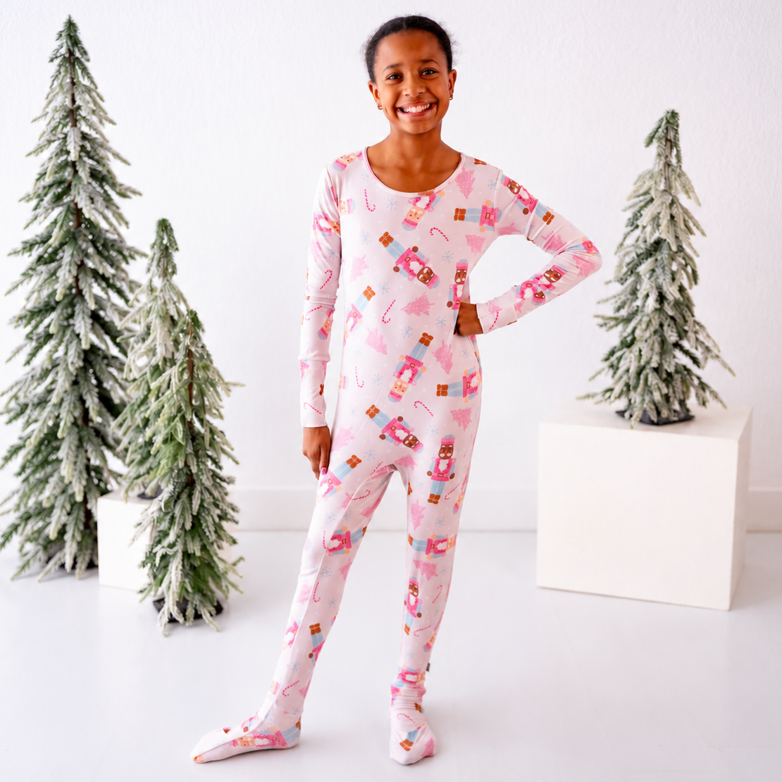 A smiling girl stands in a bright studio wearing a long-sleeve full-body pajama set with a pink holiday print. The pajamas feature illustrations of candy canes, pine cones, and wrapped gifts on a pale pink background.