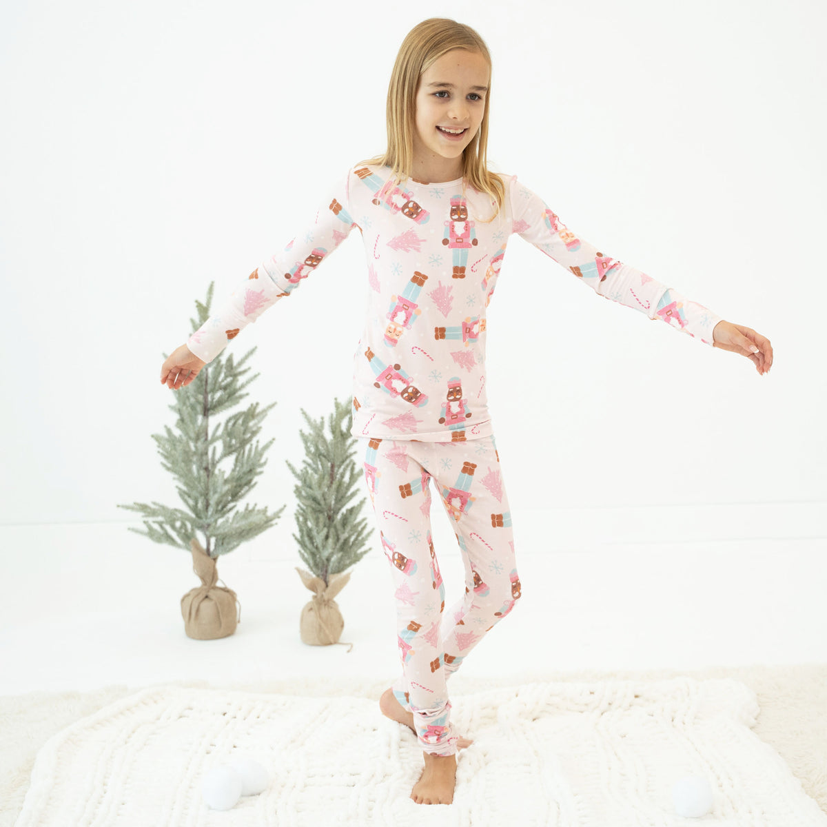 A young girl stands barefoot on a white textured blanket, wearing a light pink long-sleeve pajama set with a Christmas print of gingerbread cookies, candy canes, and snowflakes. She is posing with her arms outstretched, with two small potted pine trees in the background against a white studio backdrop.